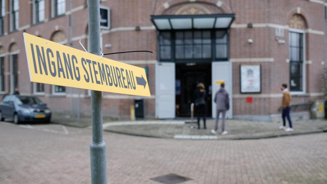 Bord met ingang stembureau en een pijl. Op de achtergrond een gebouw waar mensen naartoe lopen of voor staan.