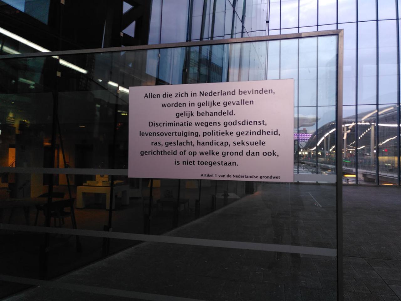 Glazen wand met daarop een bord met de tekst Allen die zich in Nederland bevinden worden in gelijke gevallen gelijk behandeld. Discriminatie wegens godsdienst, levensovertuiging, politieke gezindheid, ras, geslacht, handicap, seksuele gerichtheid of op welke grond dan ook, is niet toegestaan. Artikel 1 van de Nederlandse grondwet.