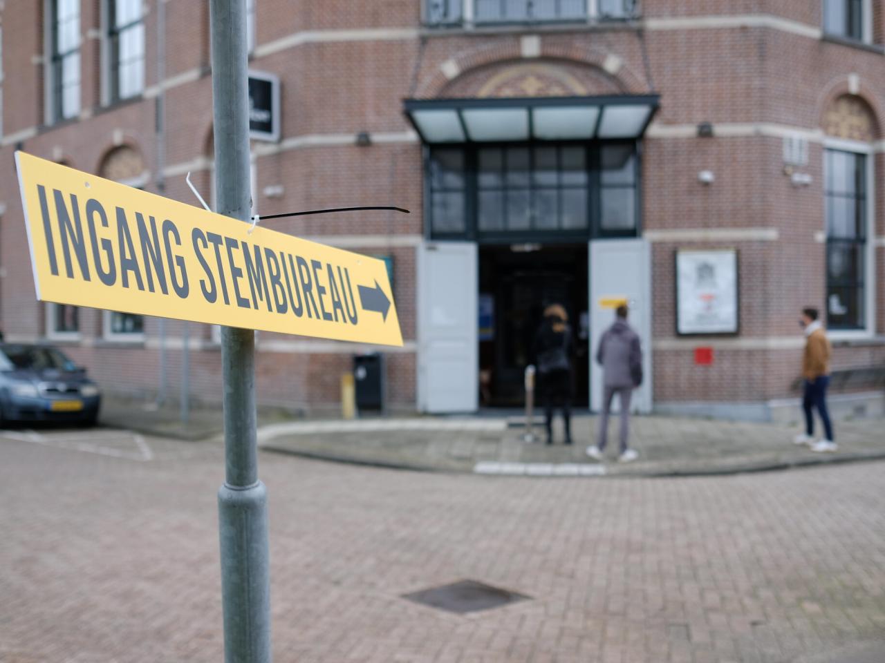 Bord met ingang stembureau en een pijl. Op de achtergrond een gebouw waar mensen naartoe lopen of voor staan.